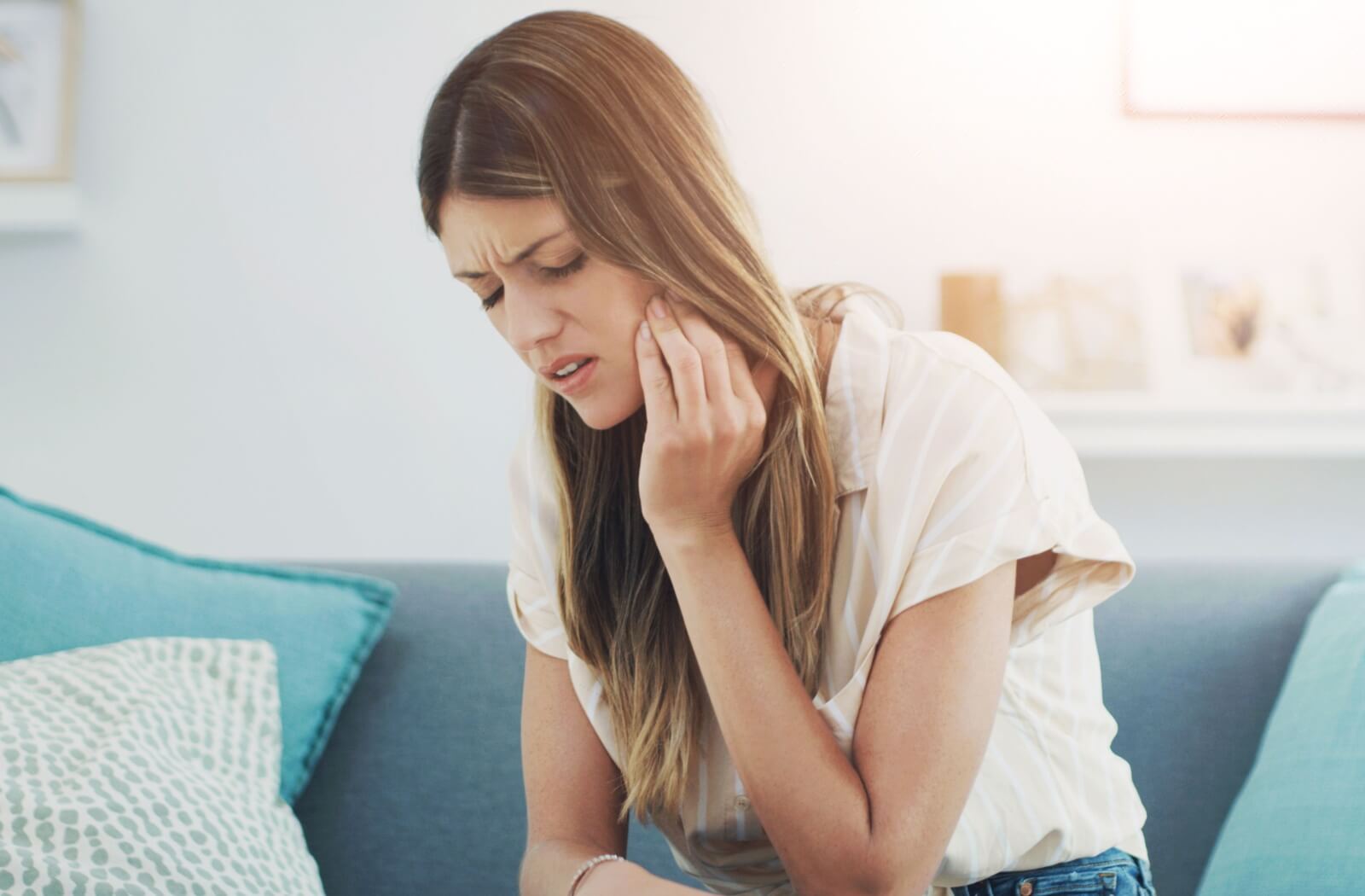 A person sits on a couch holding the side of their jaw in discomfort, suggesting wisdom tooth discomfort.