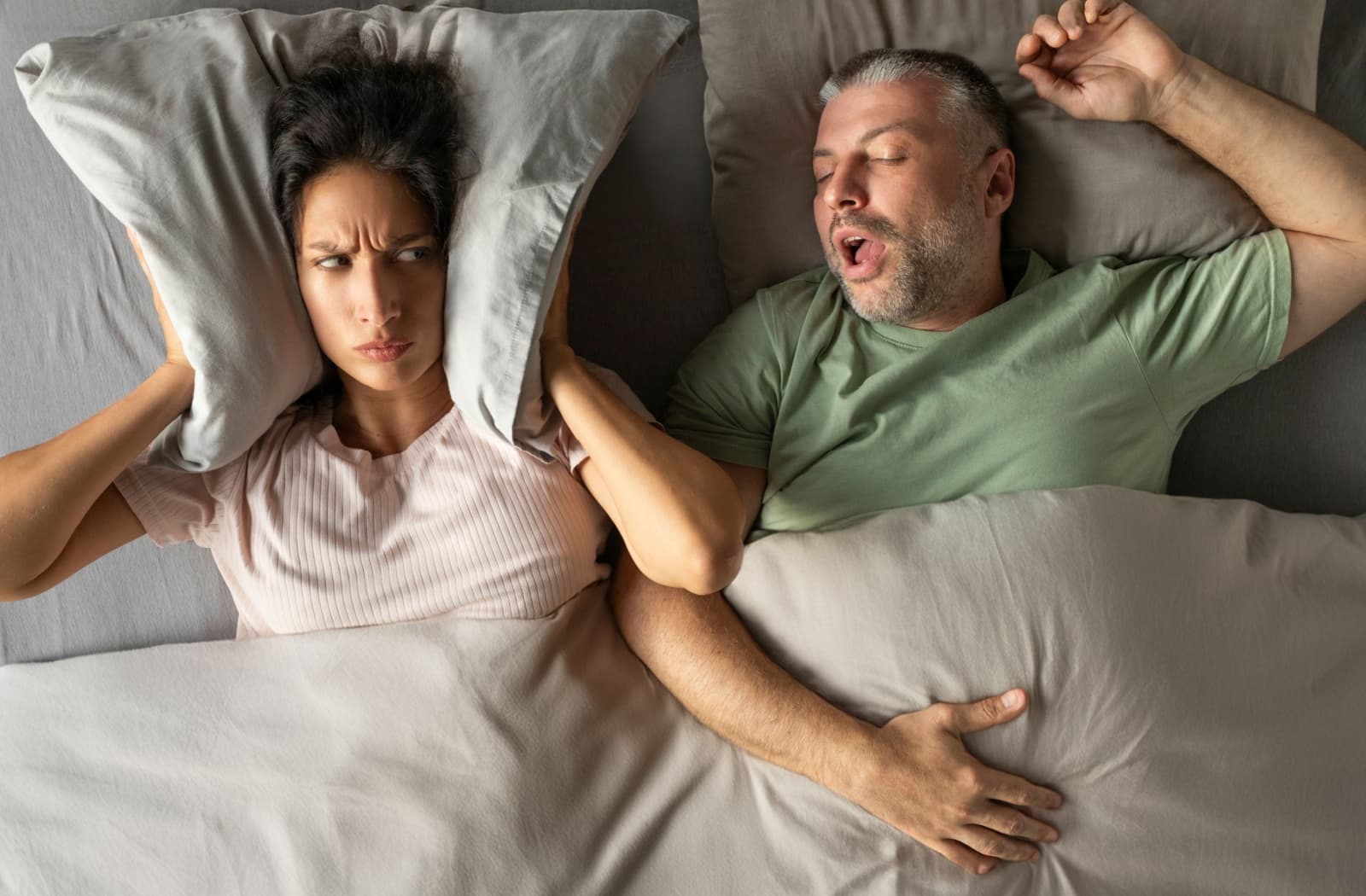 A person covering their ears with their pillow while their partner snores loudly beside them