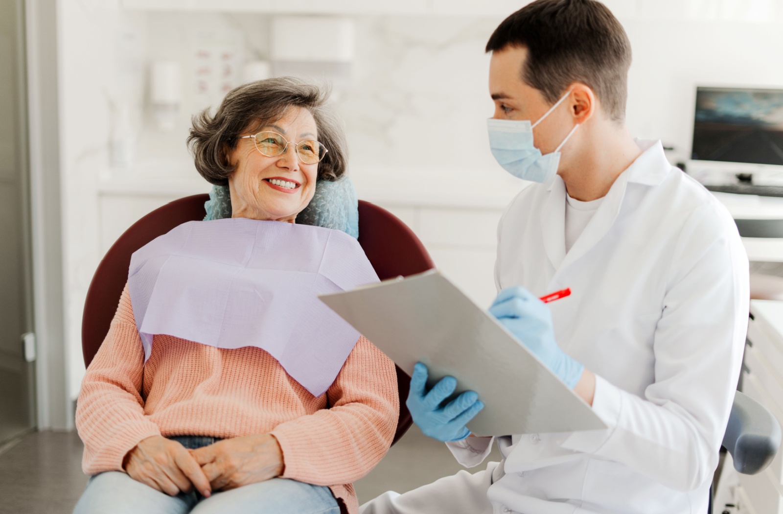 Older woman with glasses sitting in dental chair. Man in mask and white coat sits next to her writing on a clipboard.