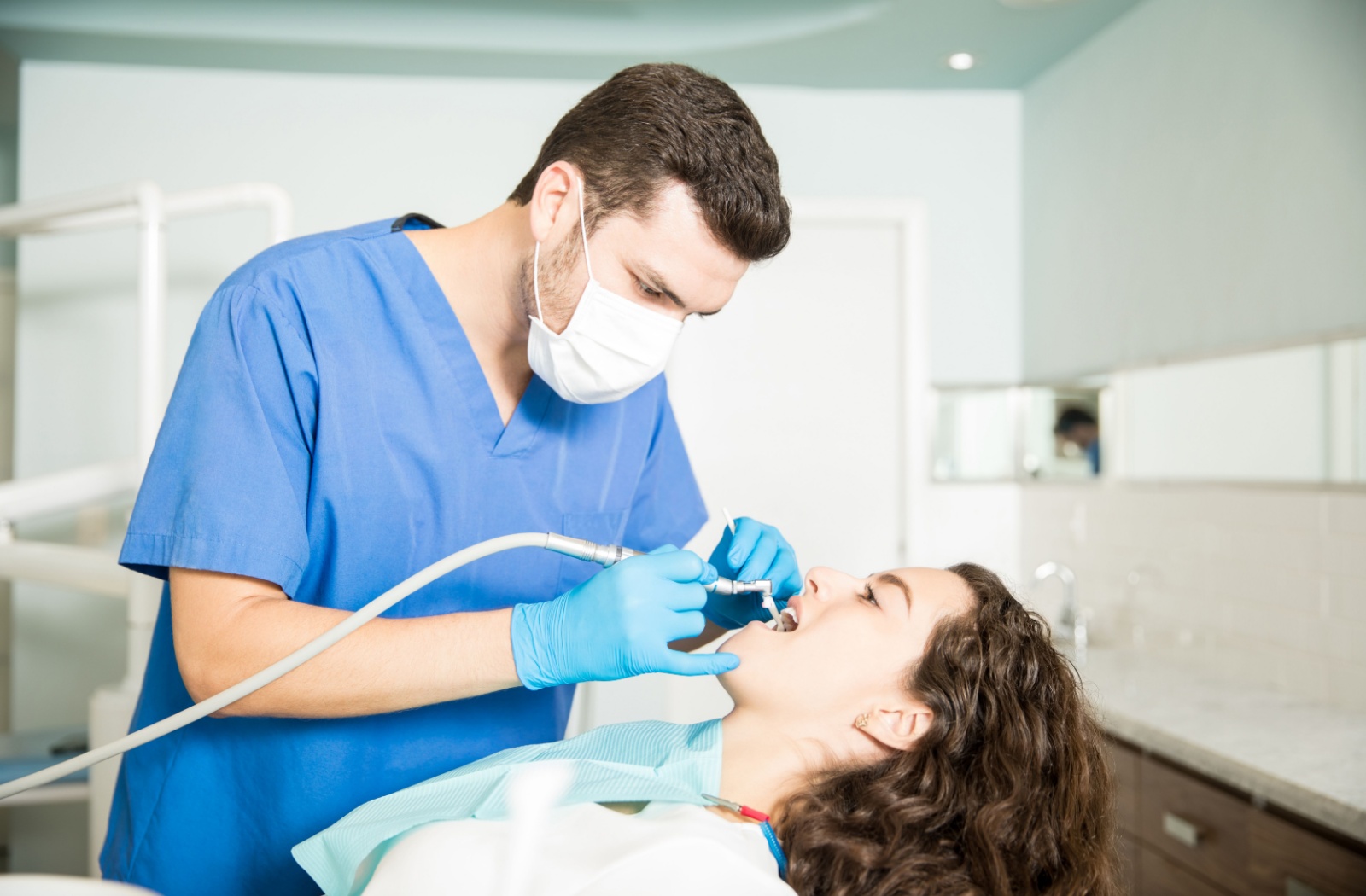 Man in blue scrubs, mask, and gloves using dental instruments to clean teeth of female patient in dental chair.