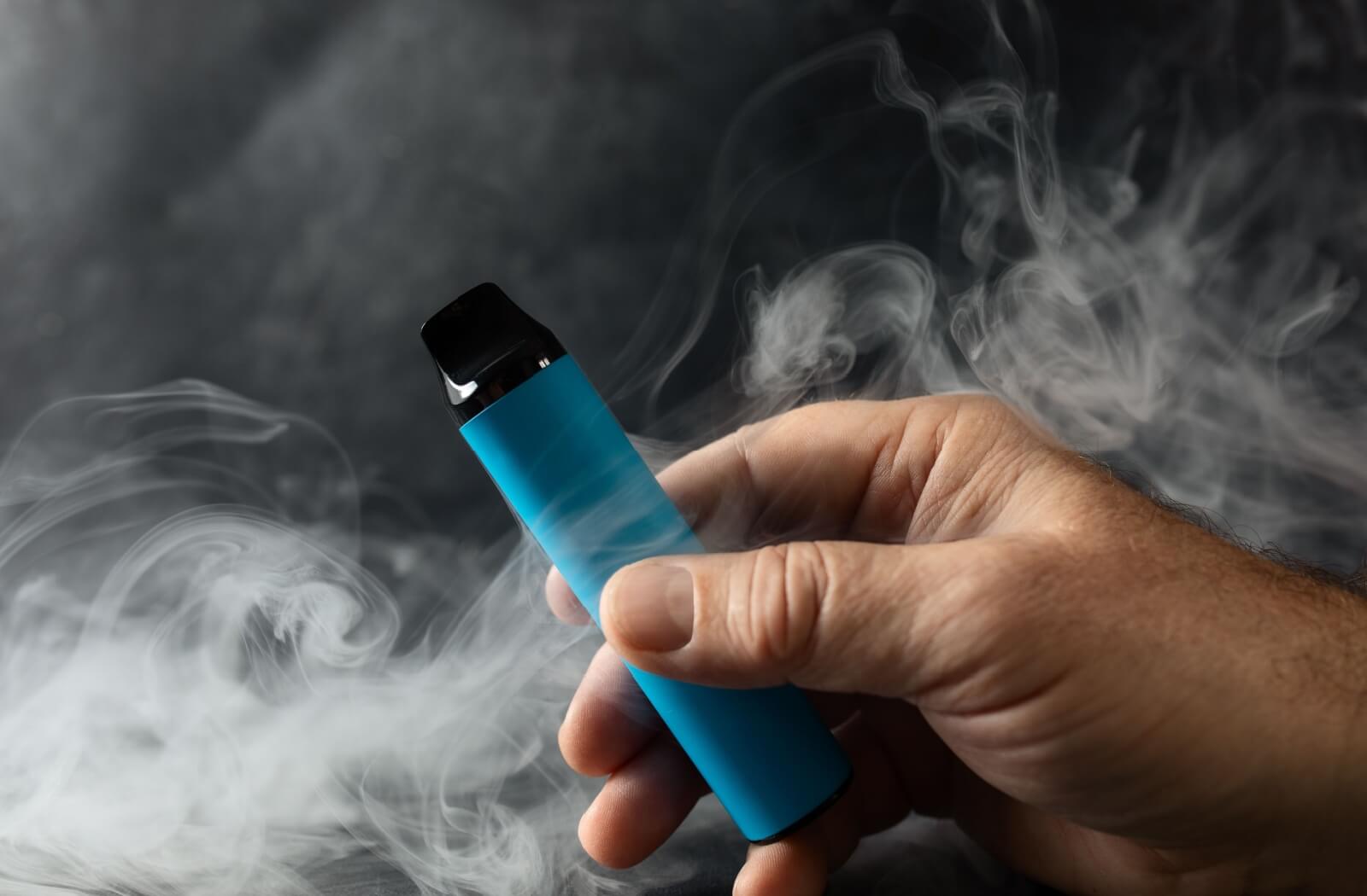 A vape device is held in a hand, with thick clouds of smoke swirling around, creating a hazy and dramatic effect.