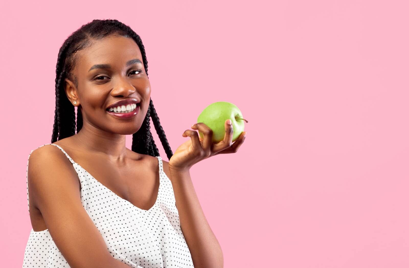 Smiling adult holding a green apple against a pink background showcasing healthy teeth and vibrant energy