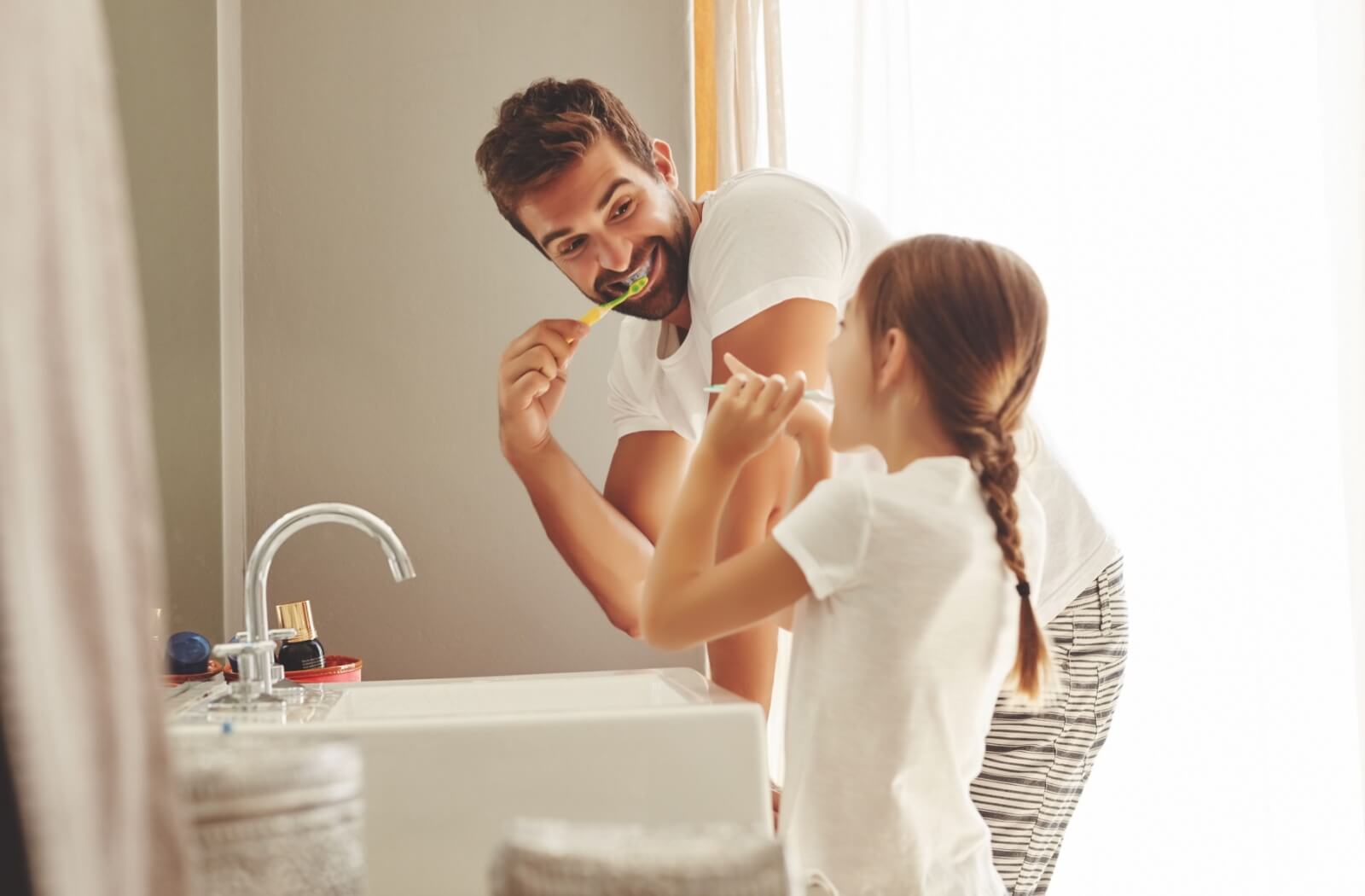 A father teaches his child about the importance of routine oral hygiene, smiling at her while he shows her how to brush his teeth.