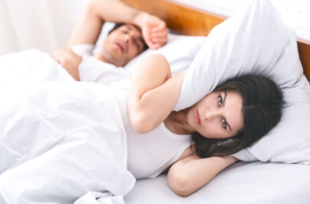 Woman in bed covering her ears with a pillow while her partner snores loudly beside her, showing the impact of sleep apnea on couples.