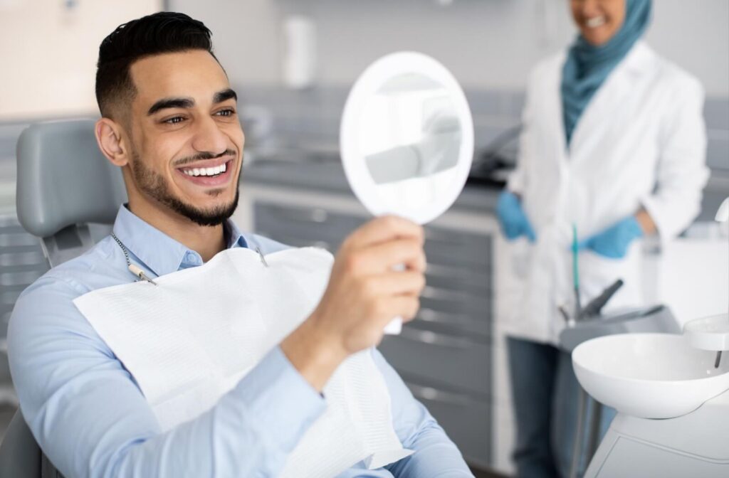 A smiling man in a dental chair holding a mirror to check his teeth while a dentist stands in the background.