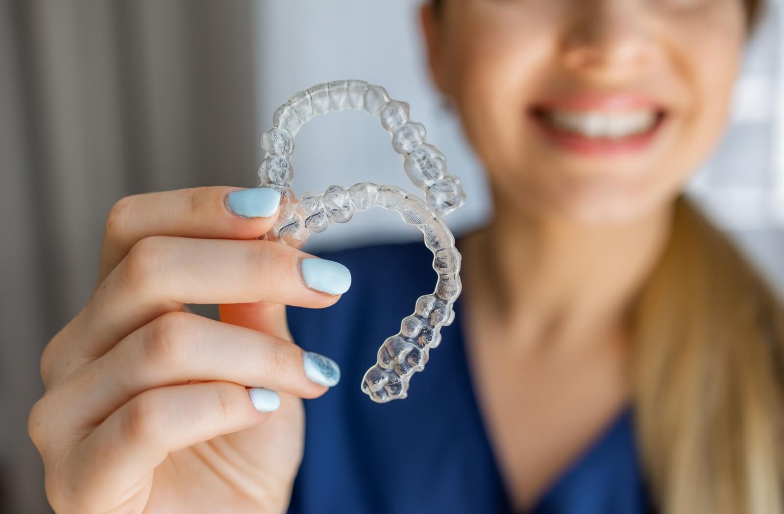 A smiling woman with light blue nail polish holding up a pair of clear Invisalign aligners, highlighting the aesthetic advantage over traditional metal braces.