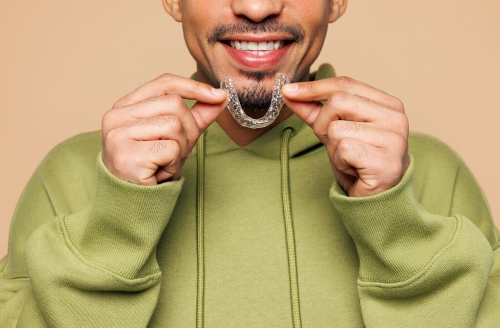 A smiling man in a green hoodie holding a clear Invisalign aligner in front of his teeth, demonstrating the discreet nature of adult orthodontic treatments.